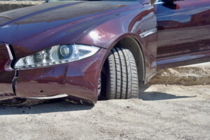 A maroon sedan is shown with a wheel stuck in a pothole. the car has taken significant damage.