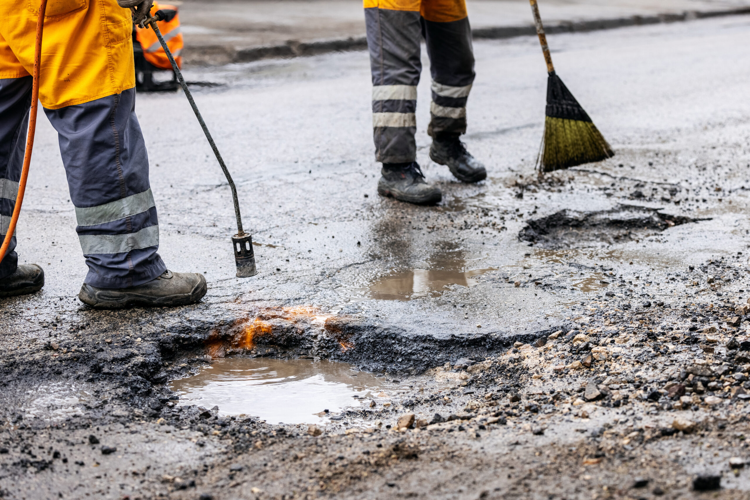 the legs of two maintenance workers are seen in the photo, but the focus is on the large pothole filled with water