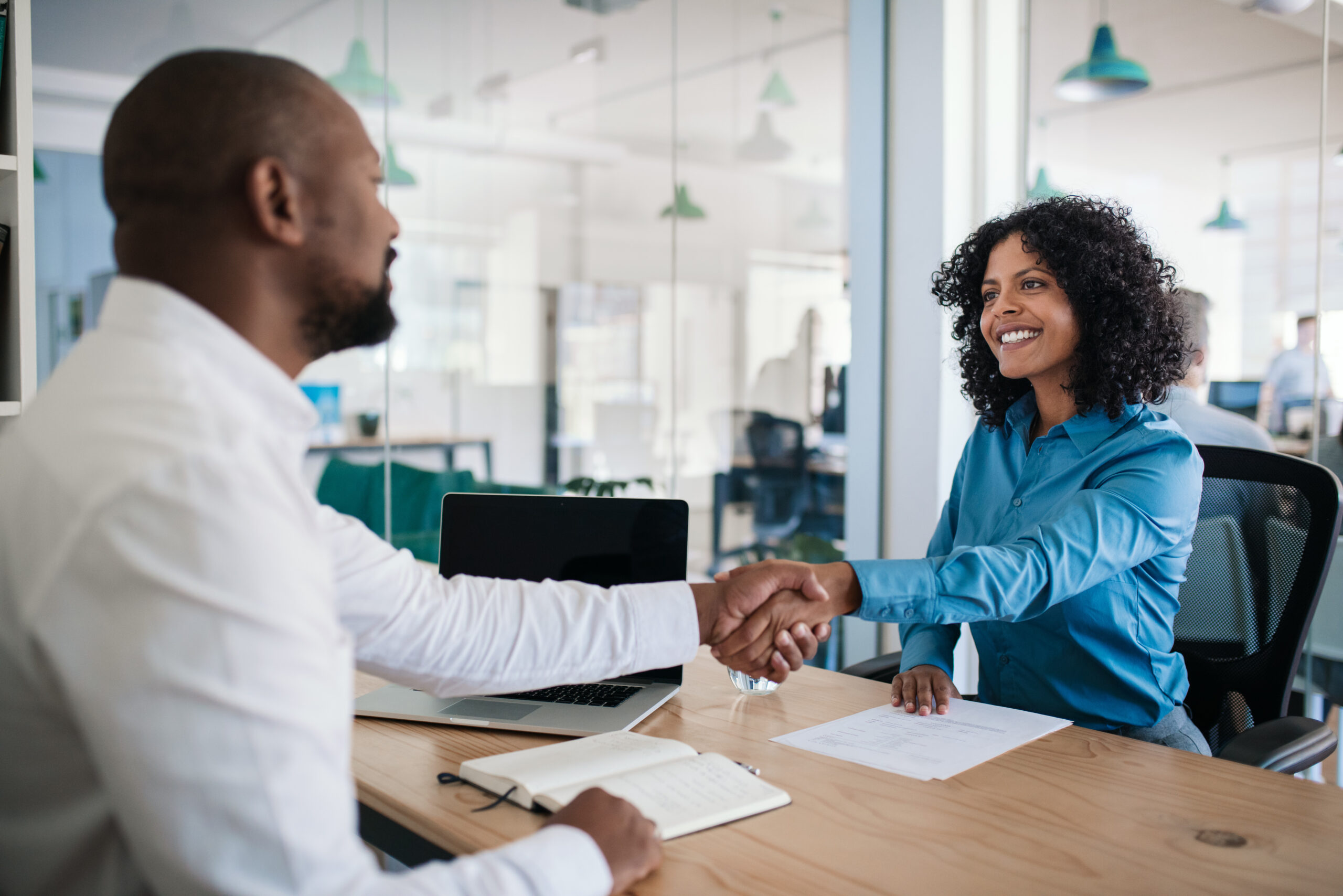 Smiling African American manager sitting at his desk in an office shaking hands with an employee after a meeting. trustee