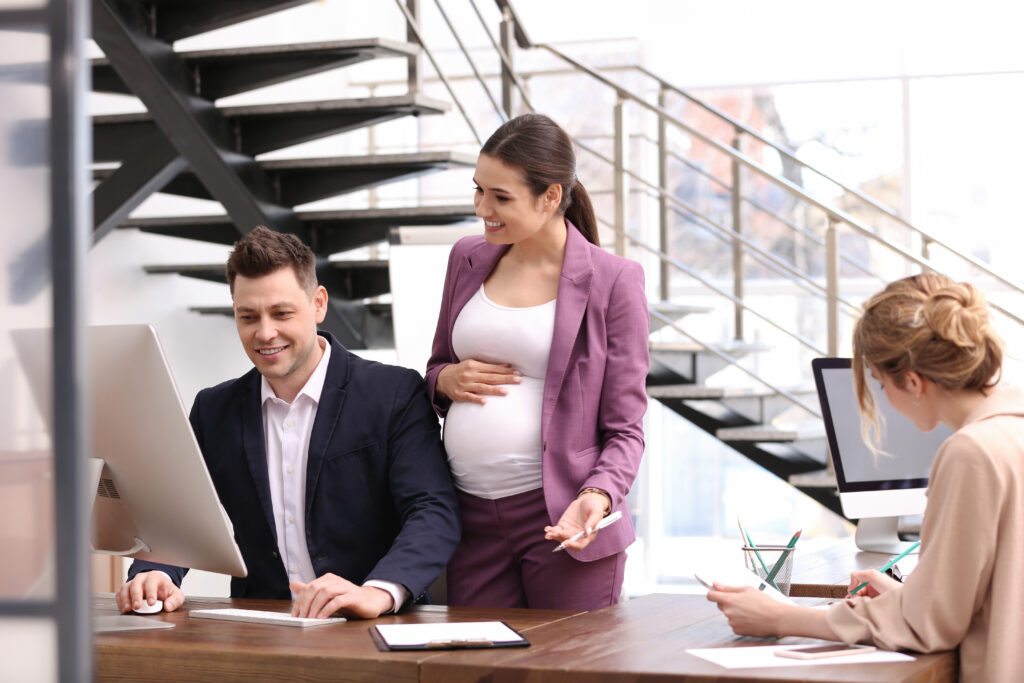 A young pregnant woman stands looking over the shoulder of her colleague while viewing something (unseen) on a computer. Modern office setting.